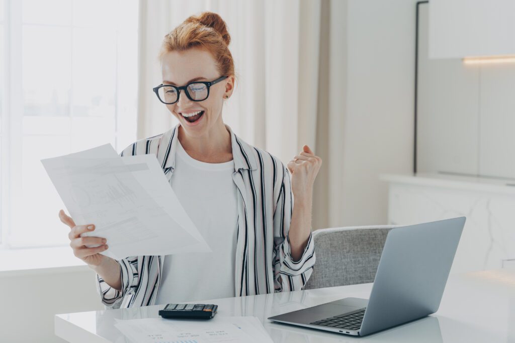 Emotional happy young ginger woman in eyewear holding paper with last mortgage payment banking notification, raising hand with clenched fist, reading document about debt consolidation.